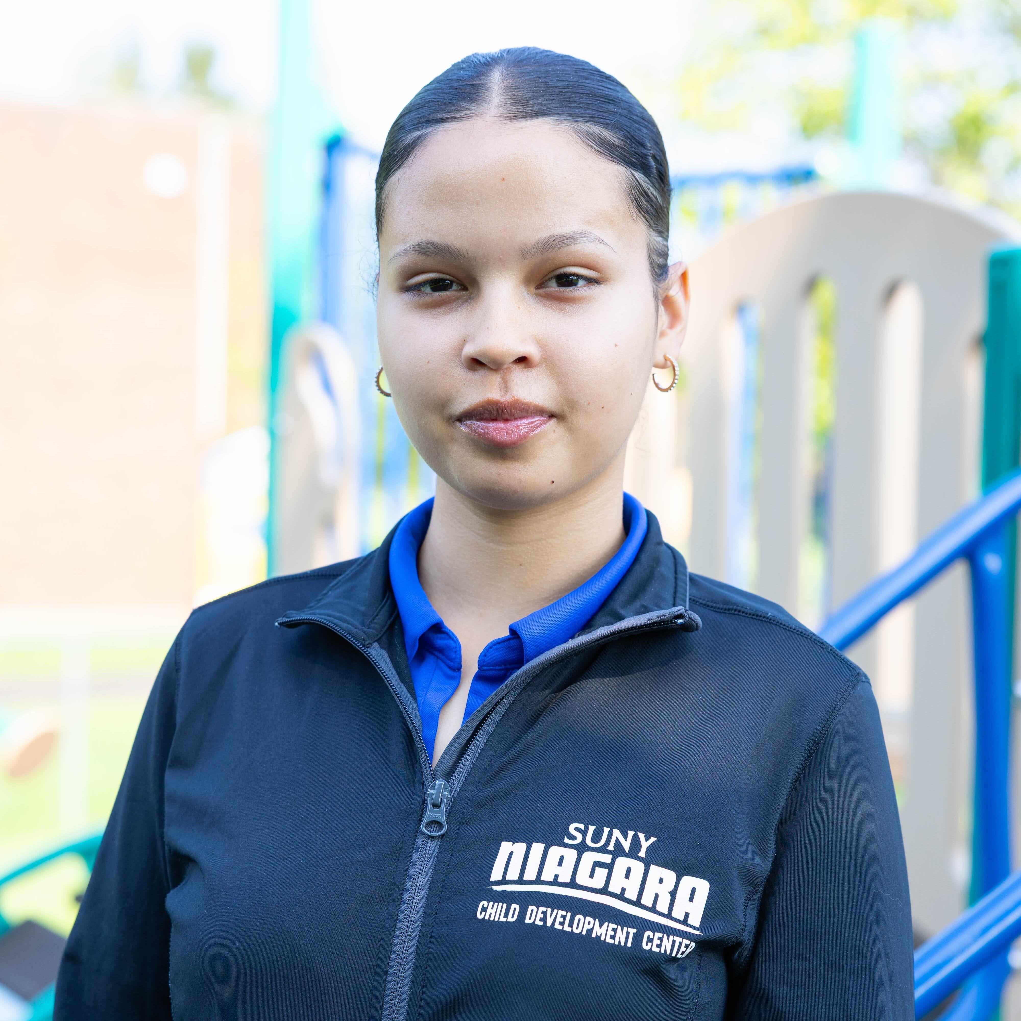 A person wearing a SUNY Niagara Child Development Center jacket stands outdoors in front of playground equipment, looking at the camera.