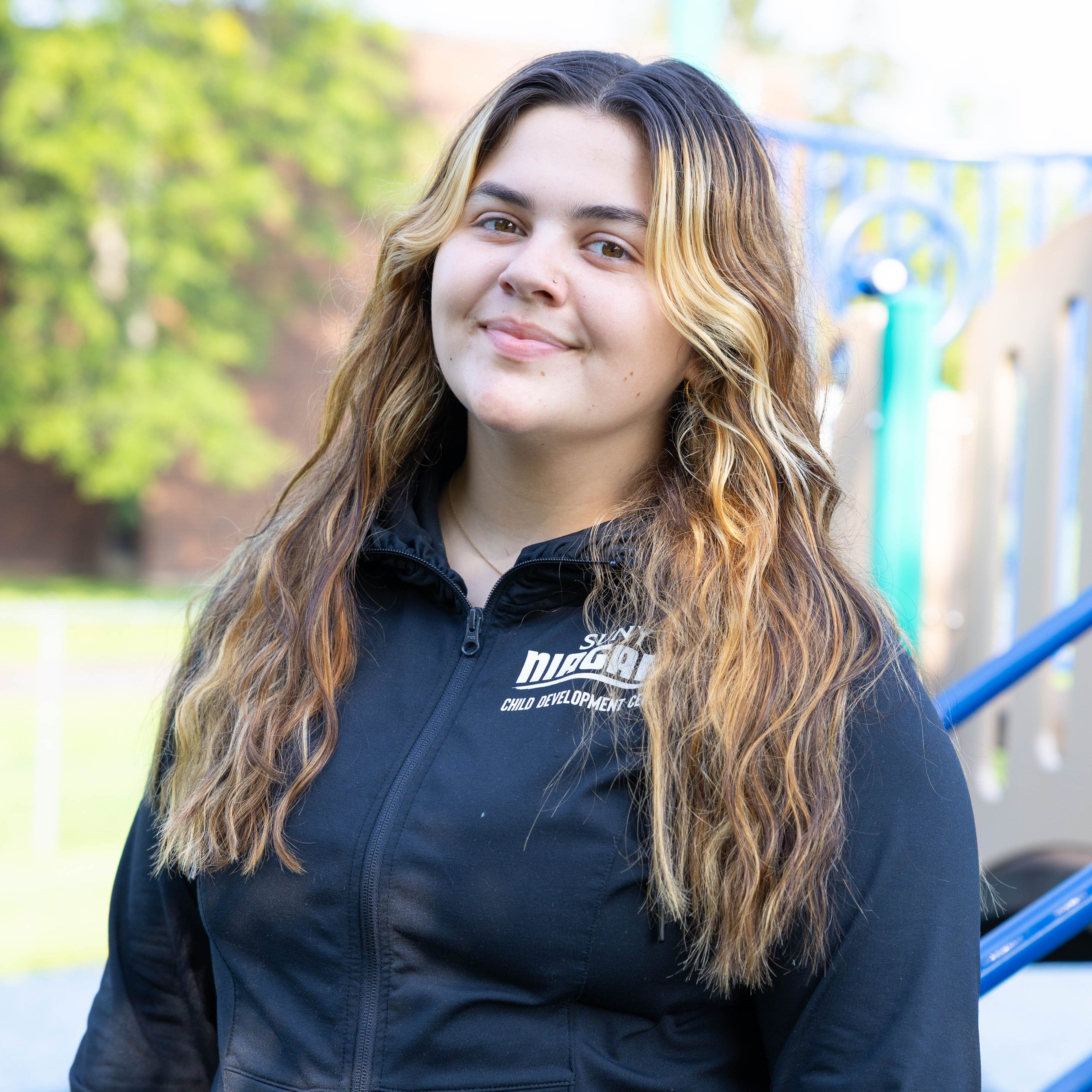 Young woman with long, wavy hair and a black zip-up jacket stands outside, smiling slightly. Playground equipment and green trees are visible in the blurred background.