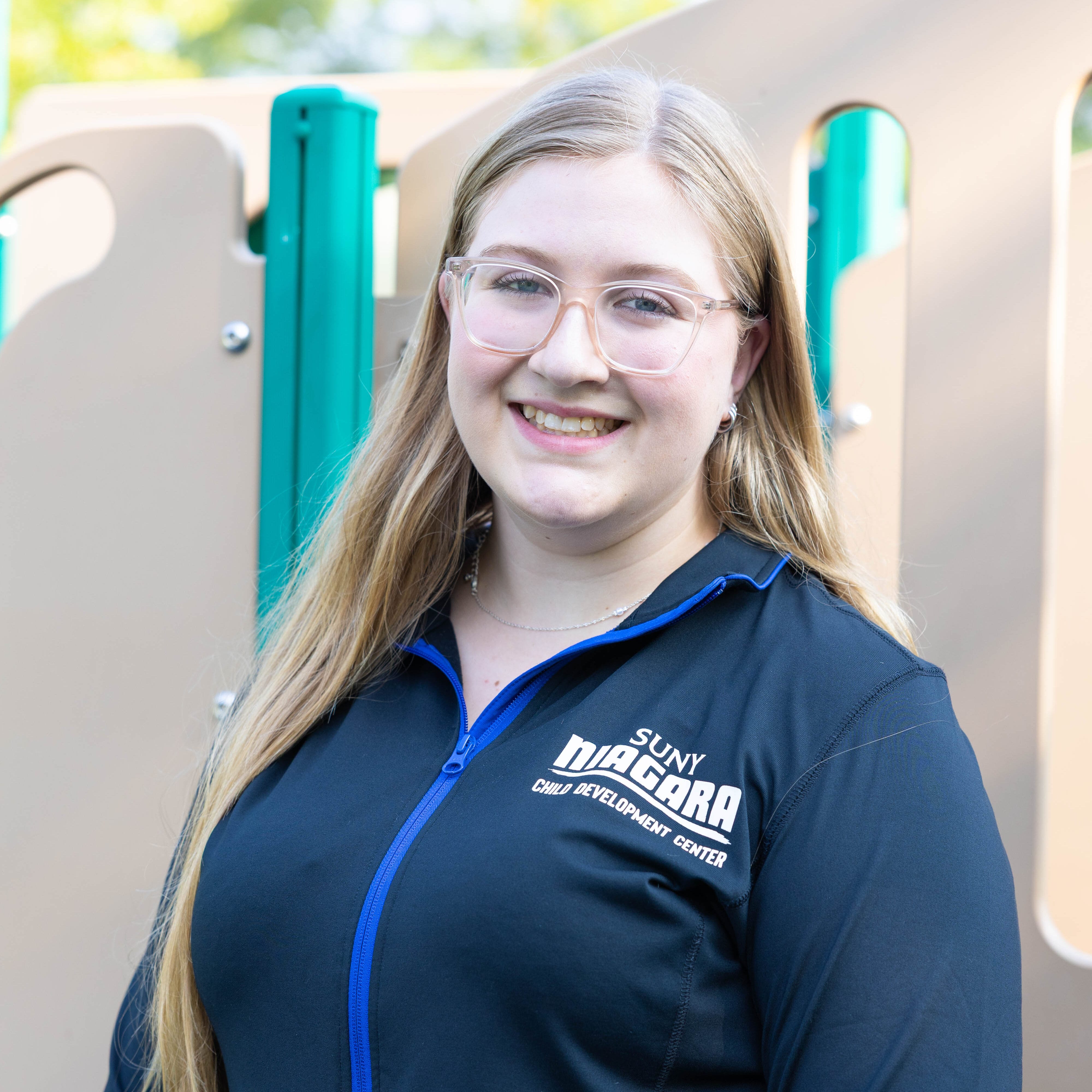 A smiling woman with long blonde hair and glasses wears a navy blue SUNY Niagara Child Development Center zip-up jacket, standing outside near tan and green playground equipment.
