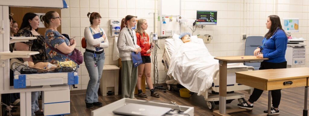 A group of students listens to an instructor in a hospital simulation room, with a medical mannequin in a hospital bed and various equipment visible.
