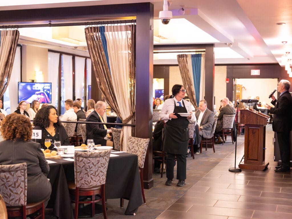 A formal event in a restaurant with seated guests, a server in an apron, and a person speaking at a podium. The room is well-lit, with tables numbered and people engaged in conversation.