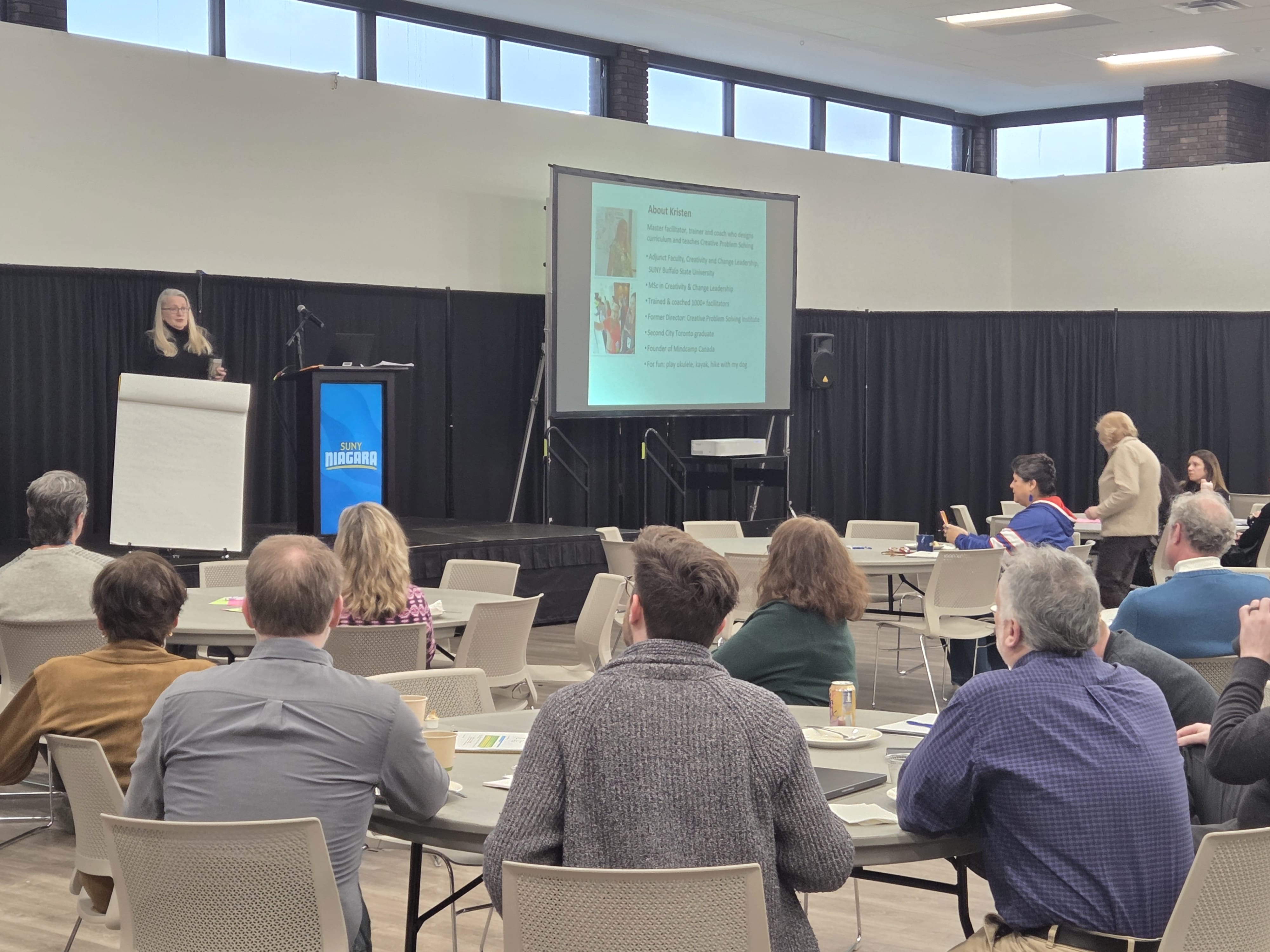 A woman stands at a podium and speaks to an audience in a conference room, embodying the spirit of One Vibrant Community. Attendees sit at tables, some taking notes, while a presentation highlighting “Discover SUNY Niagara: Two Locations” is displayed on a screen behind her.