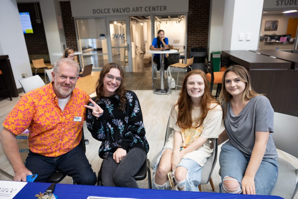Four people sit and smile at a table inside a building. The man on the left wears an orange shirt, and the three women next to him are dressed casually. Theres a sign for the Dolce Valvo Art Center in the background.