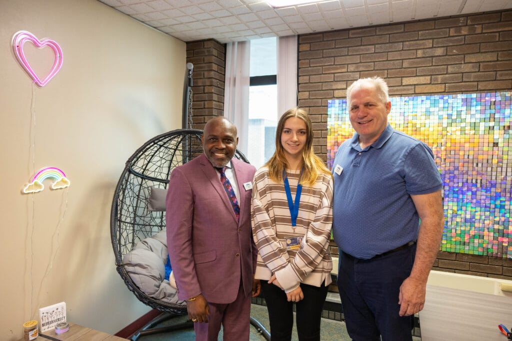 Three people stand smiling in an office with a hanging chair, neon wall art of hearts and a rainbow, and a colorful art piece on a brick wall behind them.