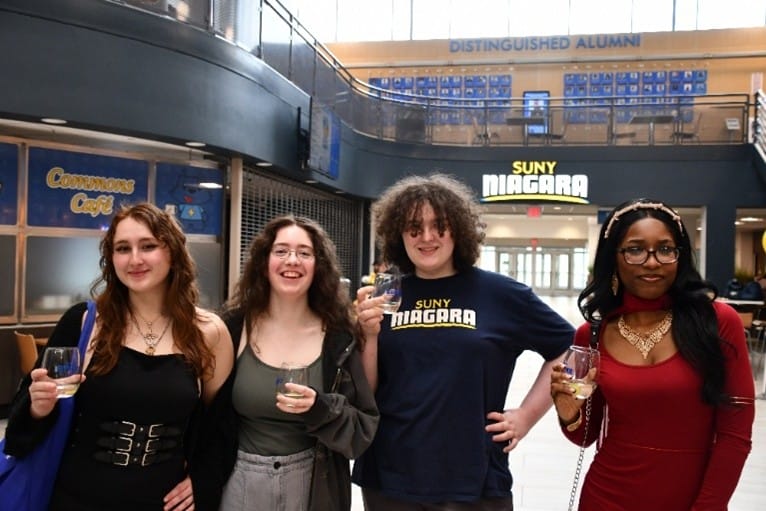 Four young adults stand indoors, smiling and holding drinks; behind them are signs for SUNY Niagara, Commons Café, and Distinguished Alumni. The setting appears to be a college campus atrium.
