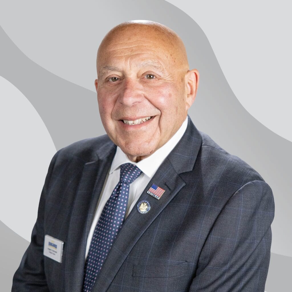 Bald man in a dark plaid suit, white shirt, and polka dot tie, smiling. He wears a US flag pin and a name badge, with a gray abstract background.