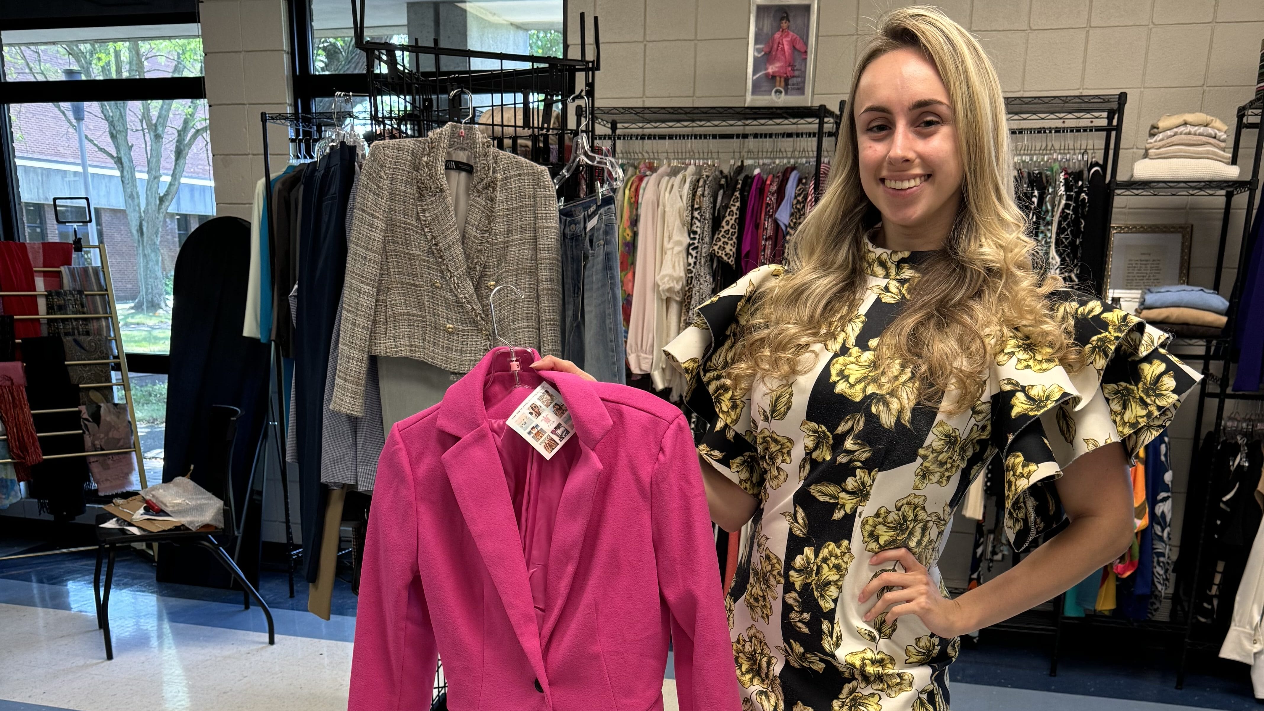 A woman in a black and white floral dress smiles and holds up a bright pink blazer on a hanger in a clothing store. Racks of clothes are visible in the background.