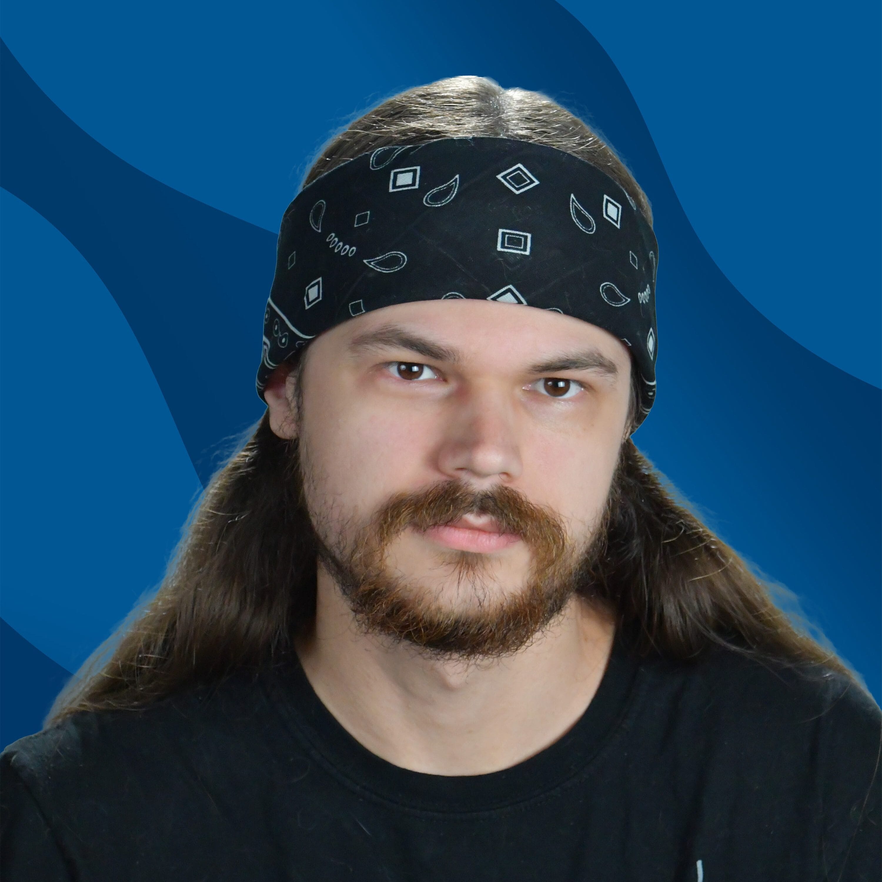A person with long hair and a beard, wearing a black bandana and a Math is Easy! Find X T-shirt, is posed in front of a blue wavy background.