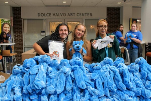 Three smiling people stand behind a table full of blue stuffed bears. They each hold up shirts or bears, and the background shows the sign Dolce Valvo Art Center. Others gather in the background.