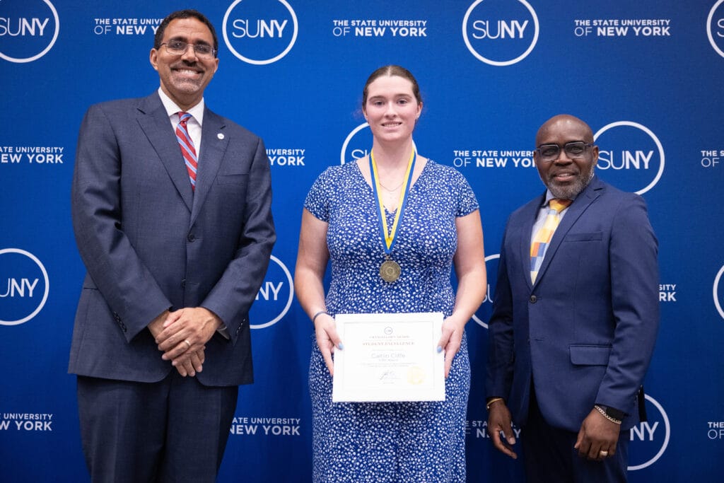 Three people stand in front of a blue SUNY backdrop. The woman in the center wears a medal and holds a certificate, while two men in suits stand on either side, all smiling at the camera.