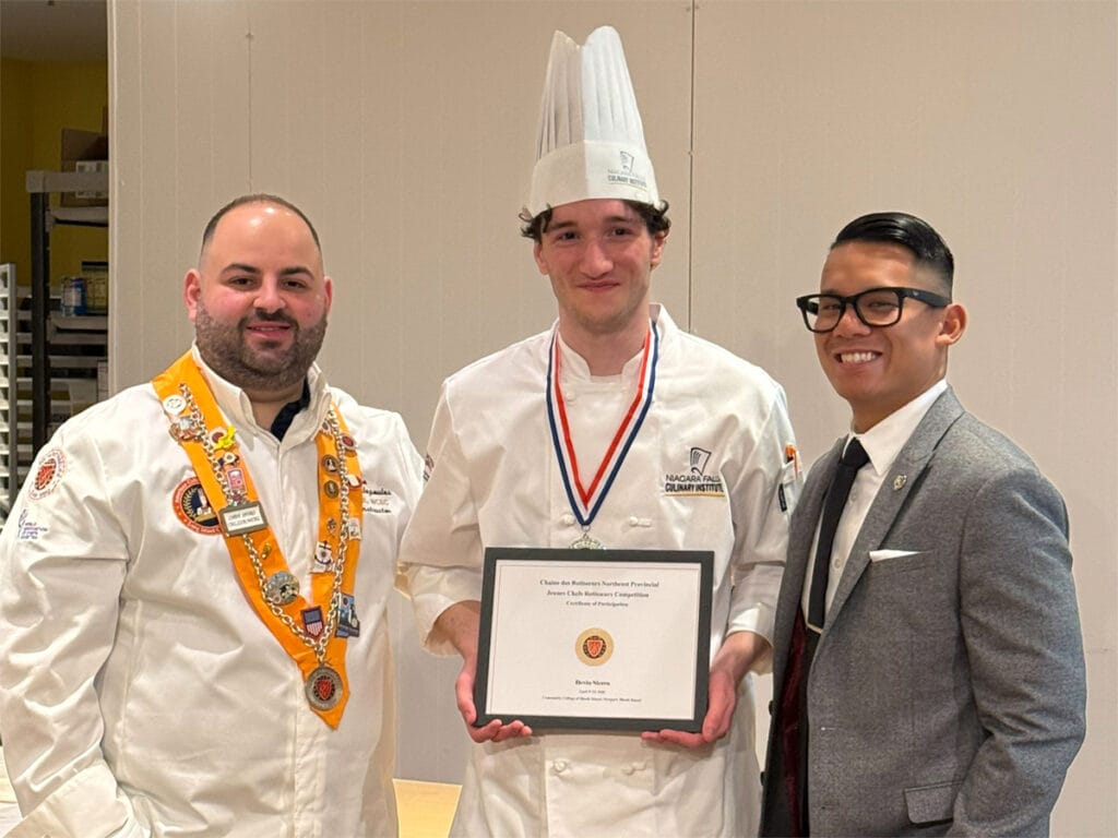 Three men pose indoors; the center man wears a chefs hat and holds a certificate, flanked by a man in a decorated chefs coat and a man in a suit and glasses.