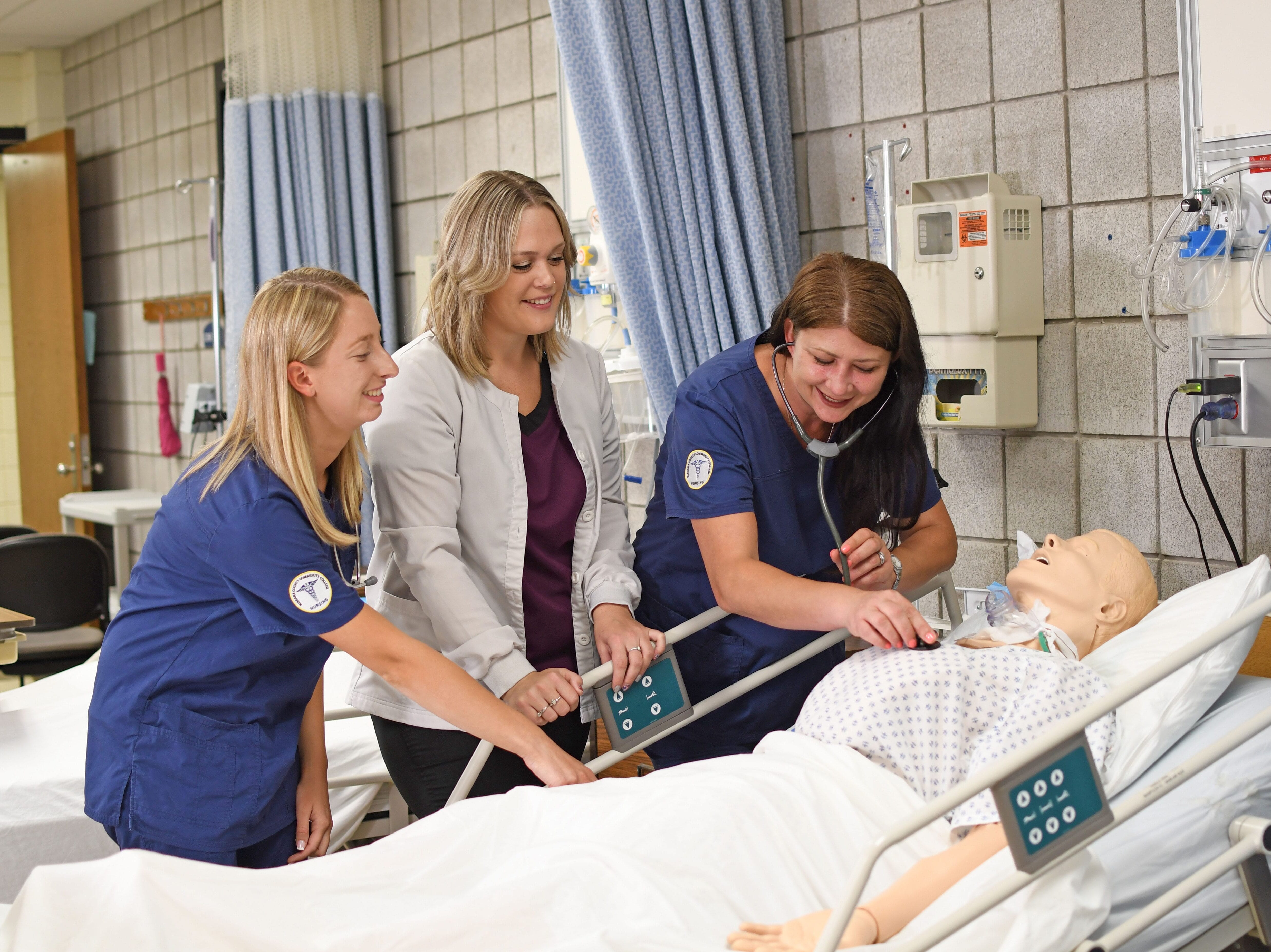 Three women, two in blue scrubs and one in a white coat, practice medical procedures on a mannequin patient lying in a hospital bed in a clinical training room.