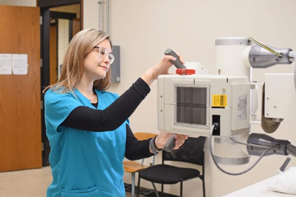 A woman in blue scrubs adjusts an X-ray machine in a medical room. She is wearing glasses and appears focused on her task.