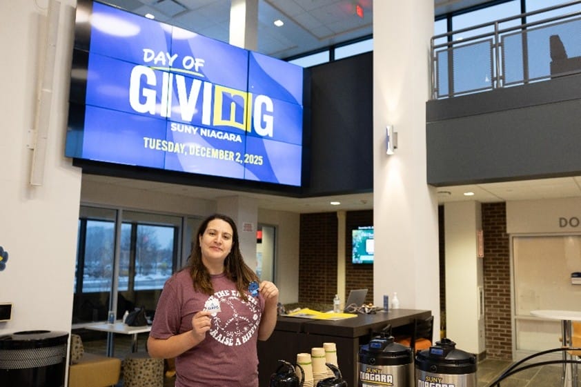 A woman stands at a table in a lobby, holding a sticker, with a Day of Giving SUNY Niagara sign and the date December 2, 2025, displayed on a large screen behind her.
