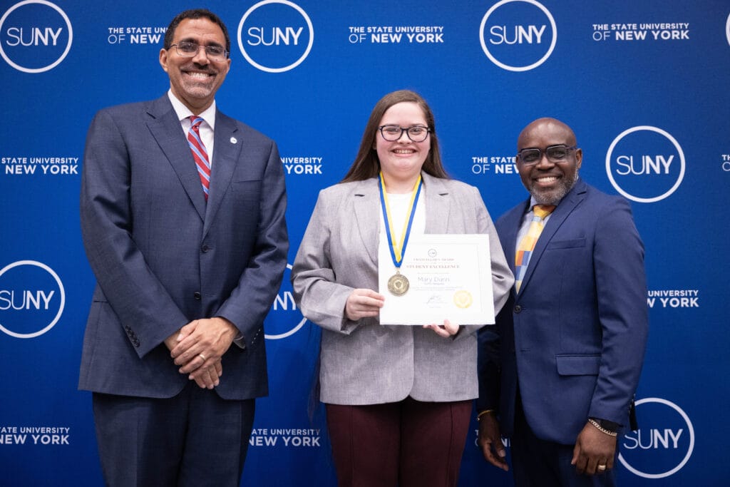 Three people stand smiling in front of a blue SUNY backdrop. The person in the center holds a certificate and wears a medal. The two others stand on either side, all dressed in business attire.