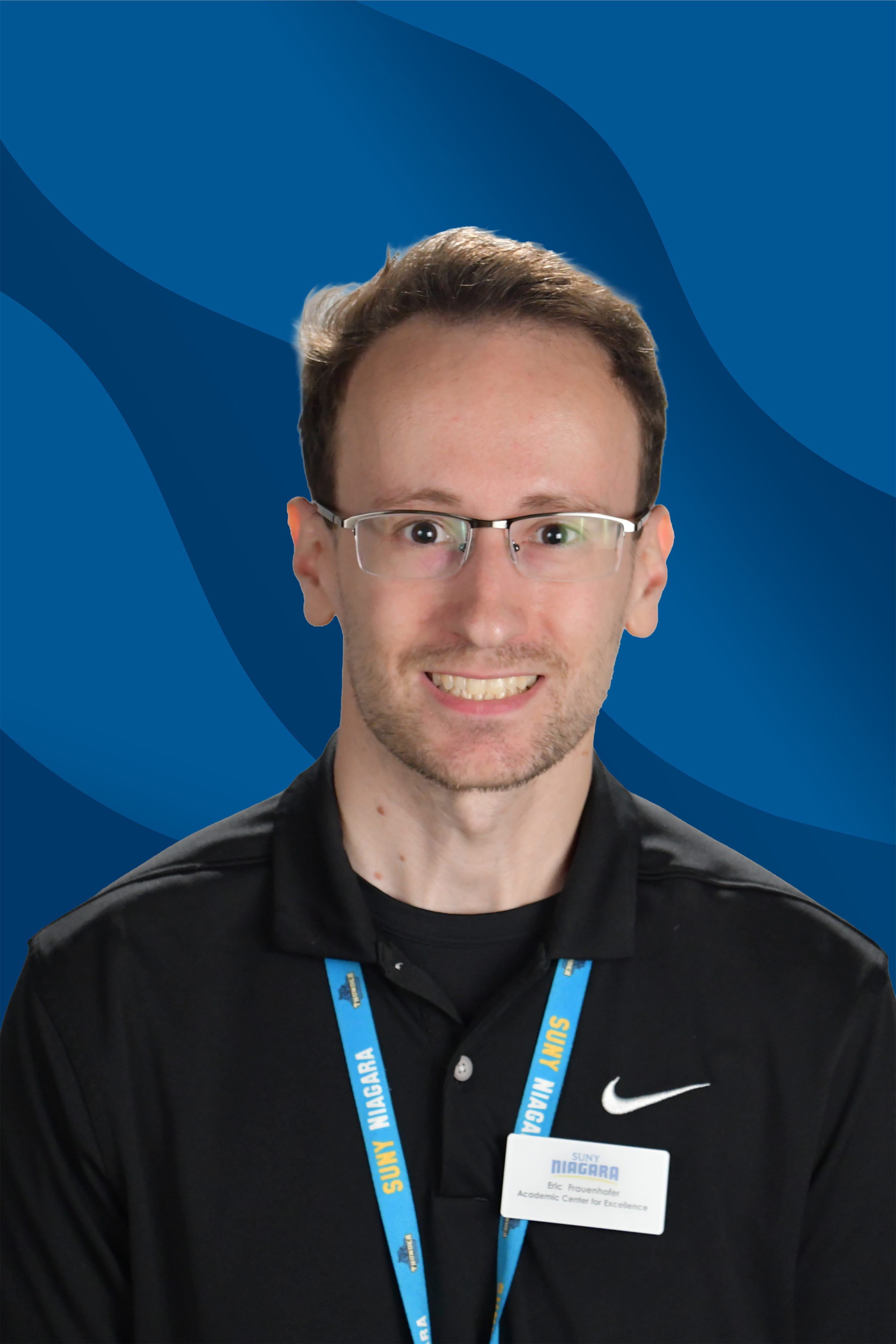 A man with short brown hair, glasses, and a black polo shirt smiles in front of a blue wavy background. He wears a blue lanyard and a name badge.