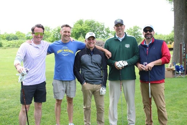 Five men stand together on a golf course, smiling at the camera. Three hold golf clubs; they are dressed casually, with greenery and trees in the background.