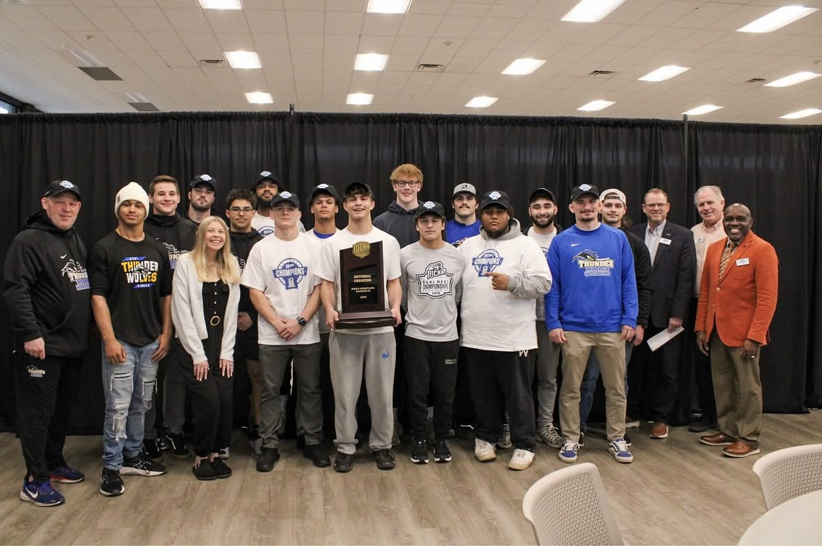 A group of young men in athletic wear and coaches stand together smiling in a large room, celebrating their wrestling teams national championship.
