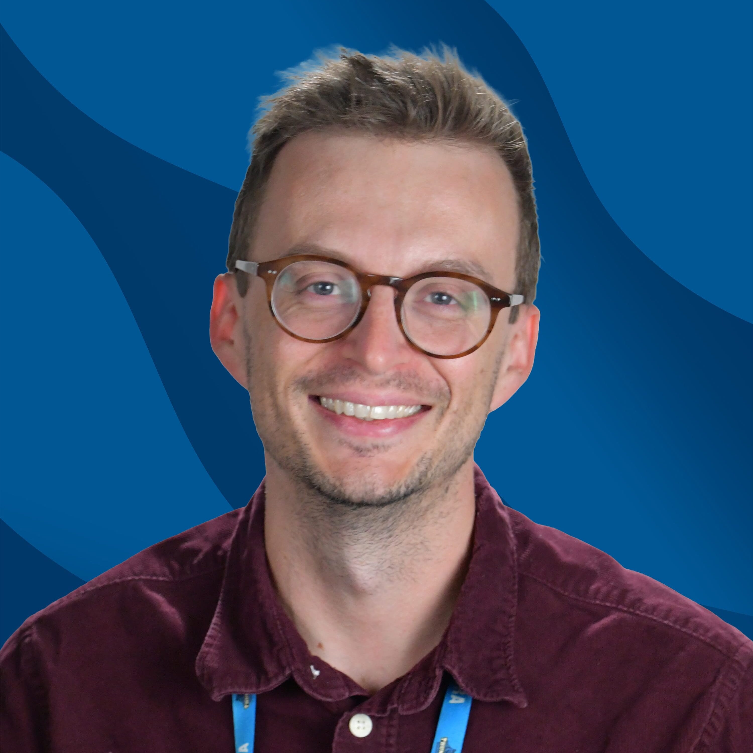A smiling person with glasses and a maroon shirt, wearing a blue SUNY Niagara lanyard, poses in front of a blue wavy background.