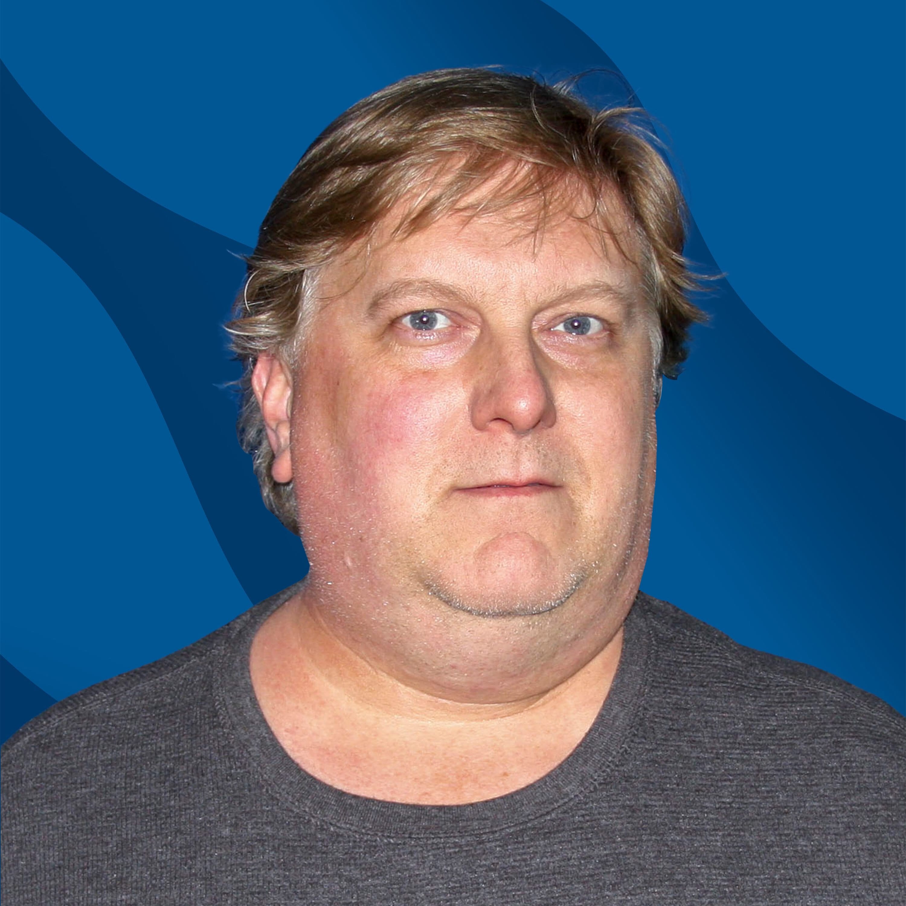 A man with light hair wearing a gray shirt stands in front of a blue background with wavy patterns, looking straight at the camera with a neutral expression.