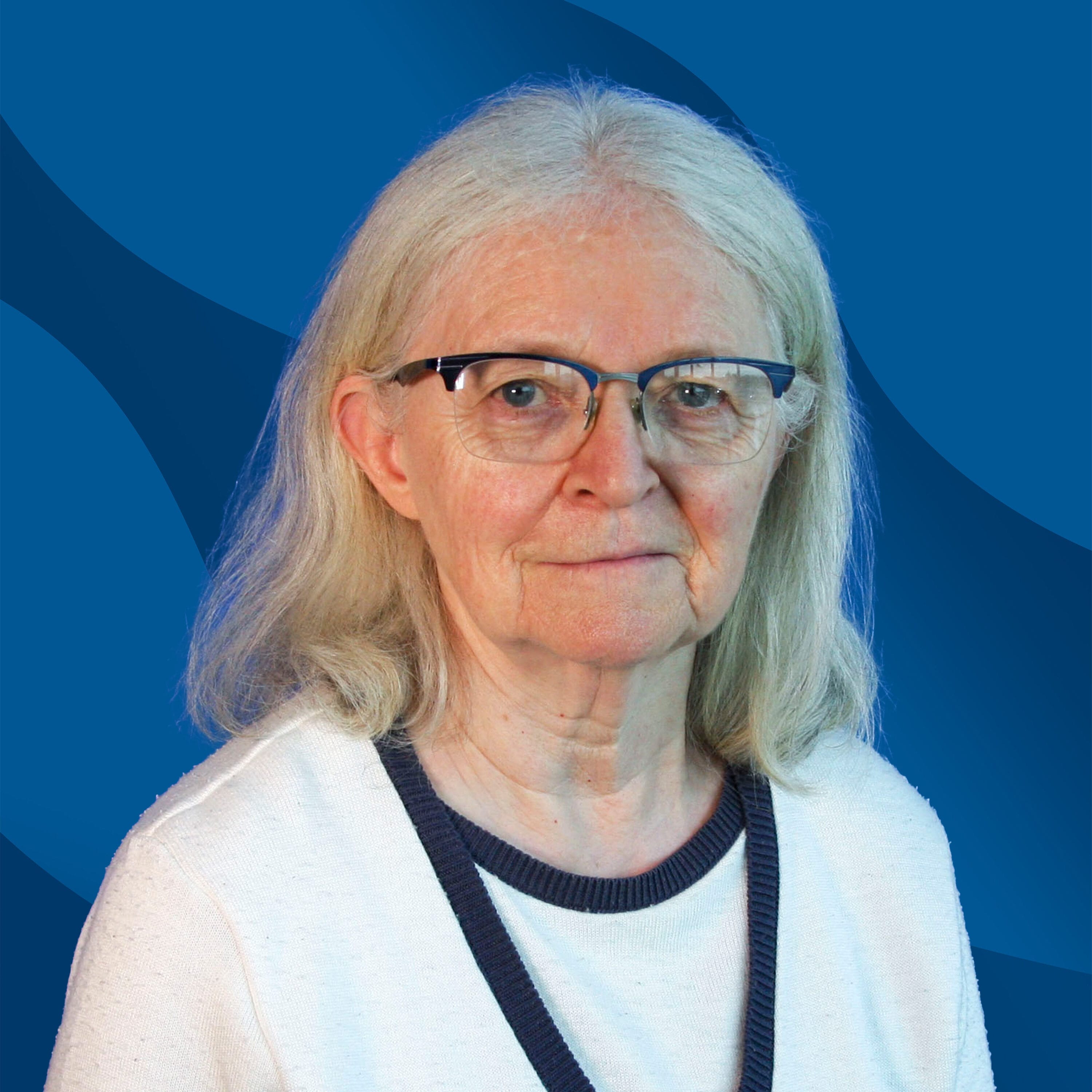 An older woman with glasses and shoulder-length gray hair wears a white cardigan with dark trim, standing in front of a blue background with wavy patterns.