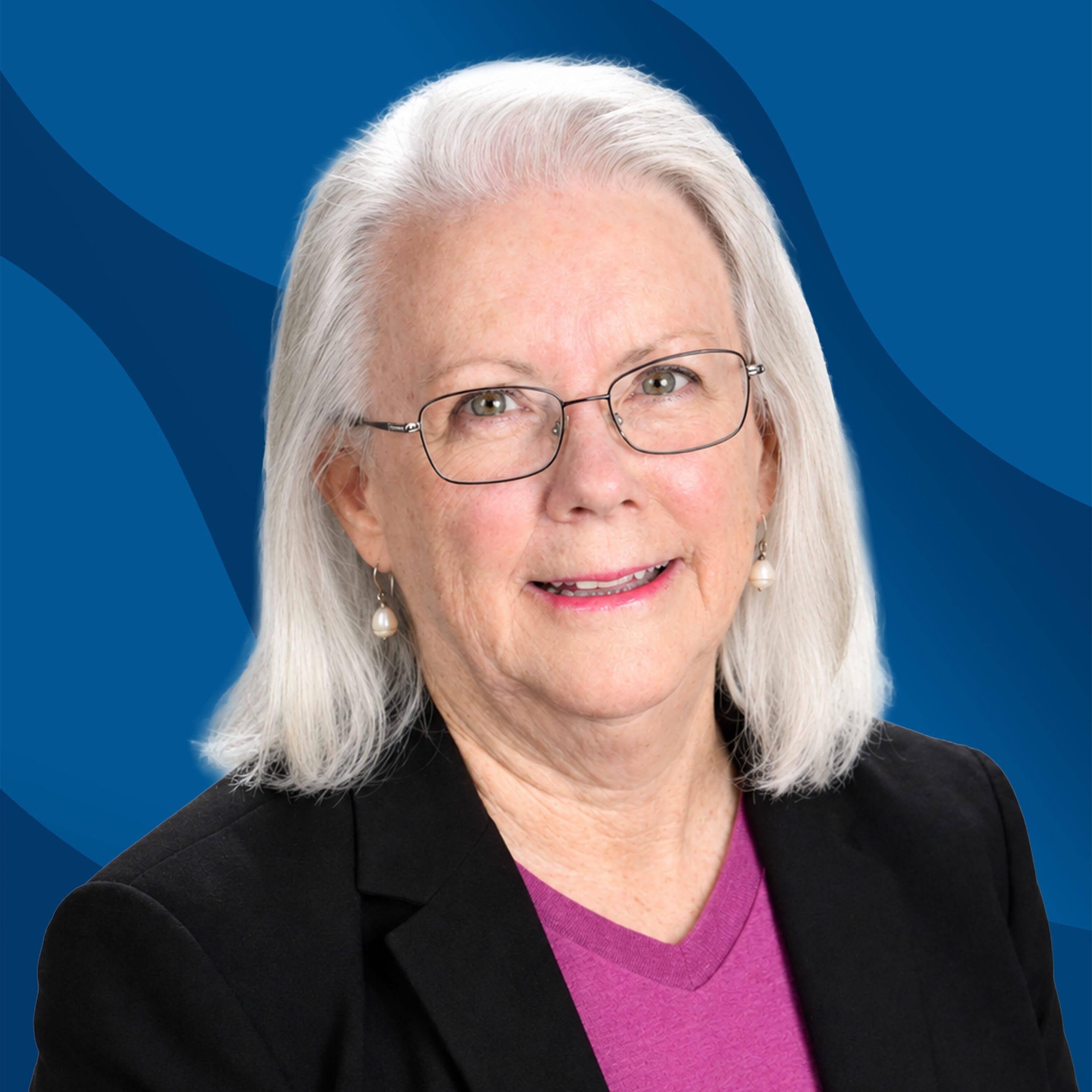 An older woman with white hair, glasses, and pearl earrings wears a black blazer over a purple top, smiling in front of a blue background with wavy patterns.