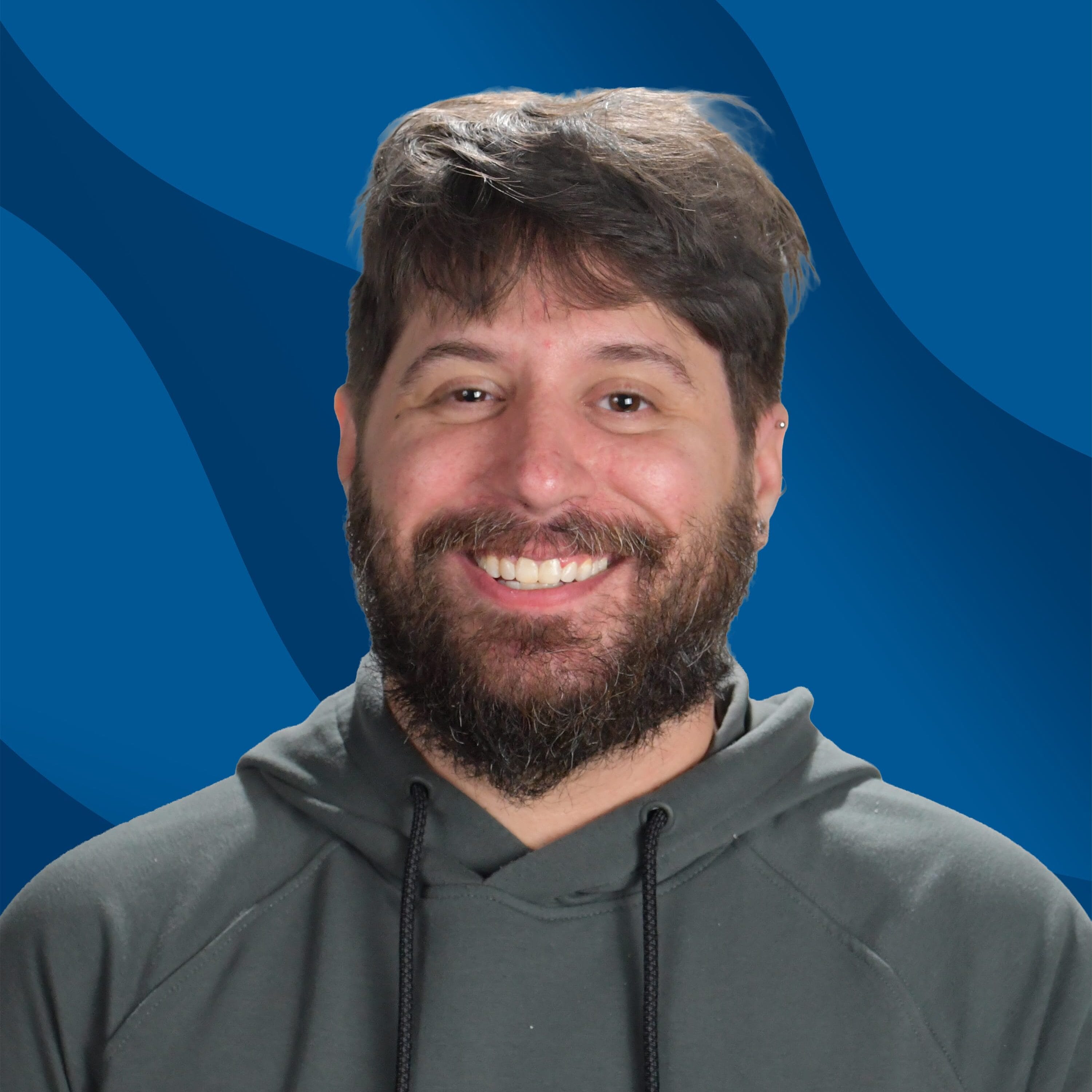 A man with brown hair and a beard, wearing a dark hoodie, smiles in front of a blue background with wavy patterns.