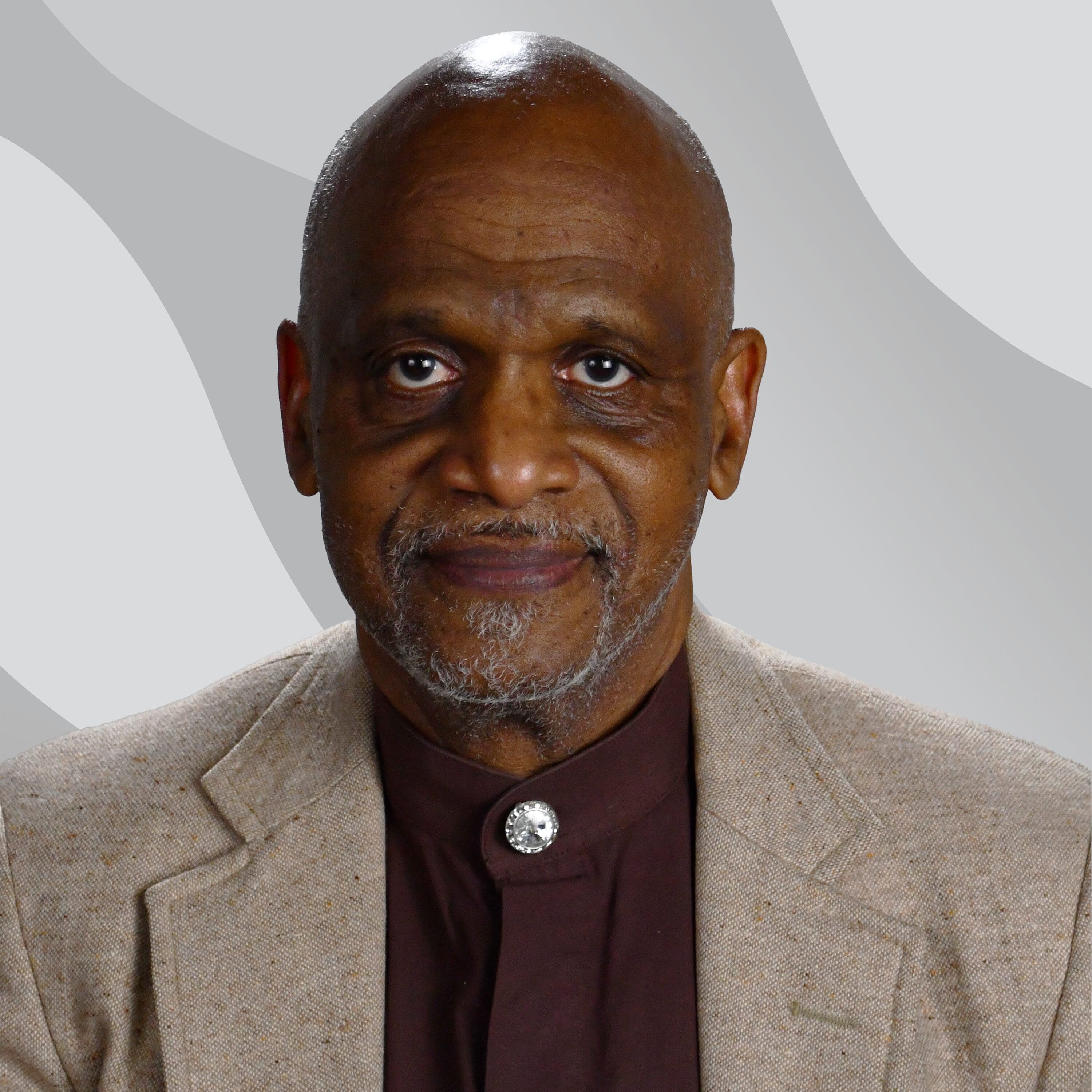 An older man with a bald head, gray beard, and mustache, wearing a tan blazer over a brown shirt with a decorative button, sits in front of a gray background with wavy patterns.