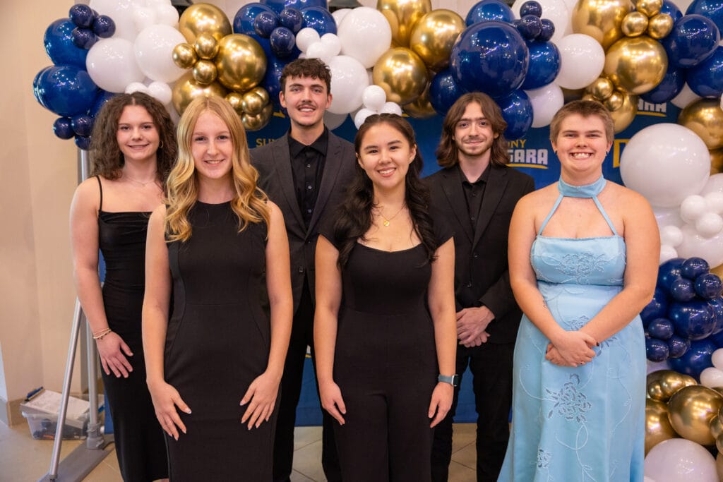 Six young adults stand in front of a backdrop decorated with blue, white, and gold balloons. Five wear black outfits, and one wears a light blue dress. They are smiling and posing for a group photo.