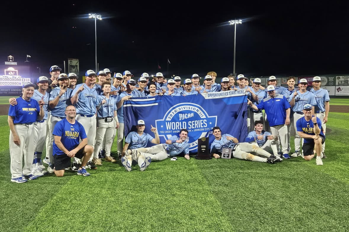 SUNY Niagara baseball team poses on a field at night, celebrating their NJCAA Division III National Championship win, with players and staff surrounding a WORLD SERIES banner.