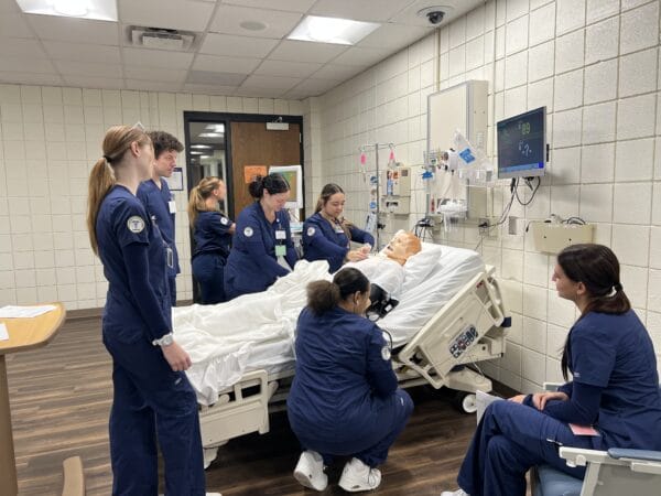 A group of nursing students in blue scrubs practice on a medical mannequin in a hospital room, observing monitors and taking notes.