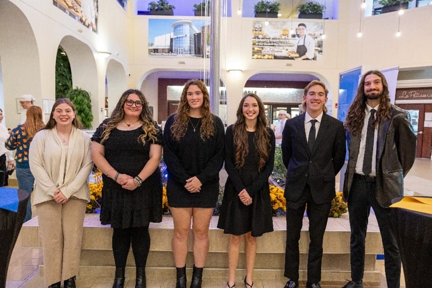 Six young adults stand indoors, dressed in formal attire, smiling at the camera. There are flowers in front of them and banners overhead in a large, bright atrium with plants and shopfronts visible in the background.