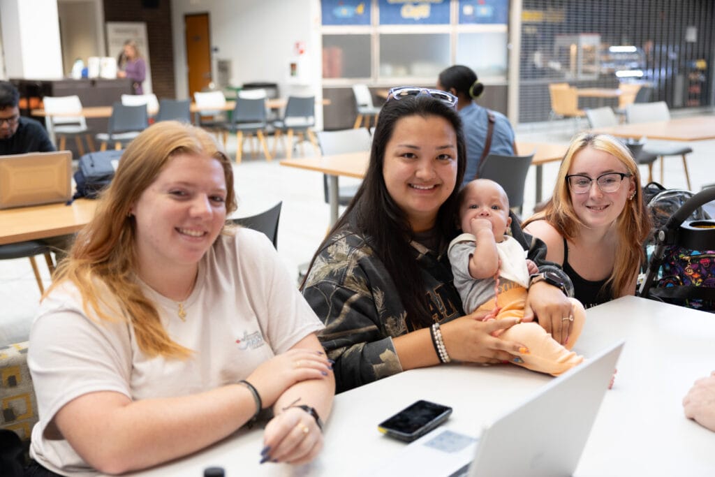 Three young women smile at a table, one holding a baby. They are seated in a casual indoor setting with people and tables in the background.
