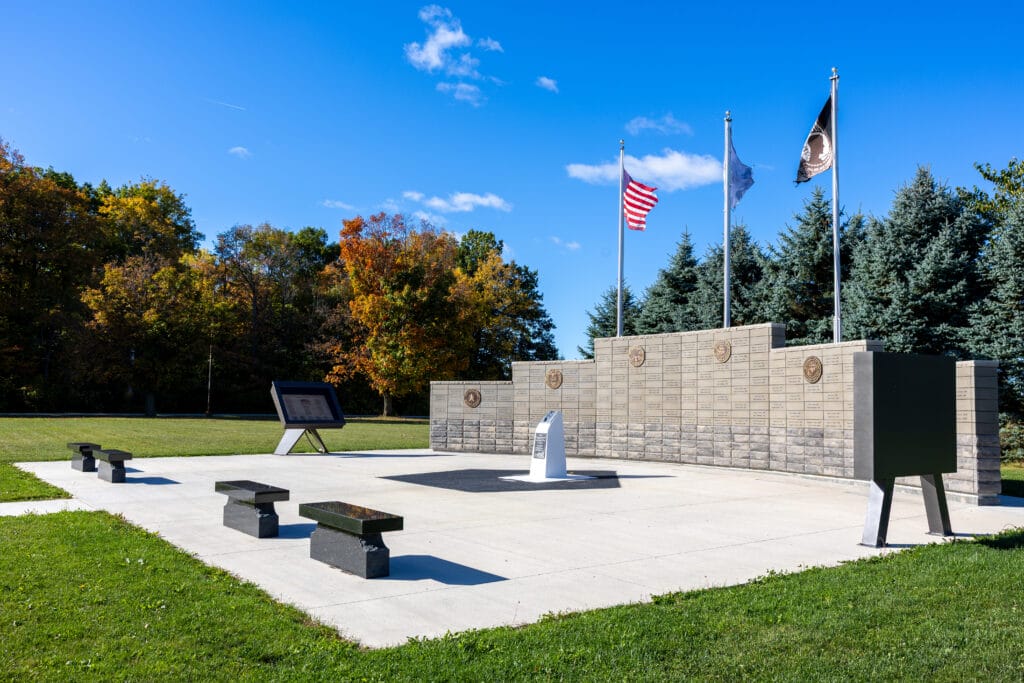 Outdoor memorial with stone wall, plaques, three flags, benches, and informational signs, surrounded by grass and trees with autumn foliage under a bright blue sky.