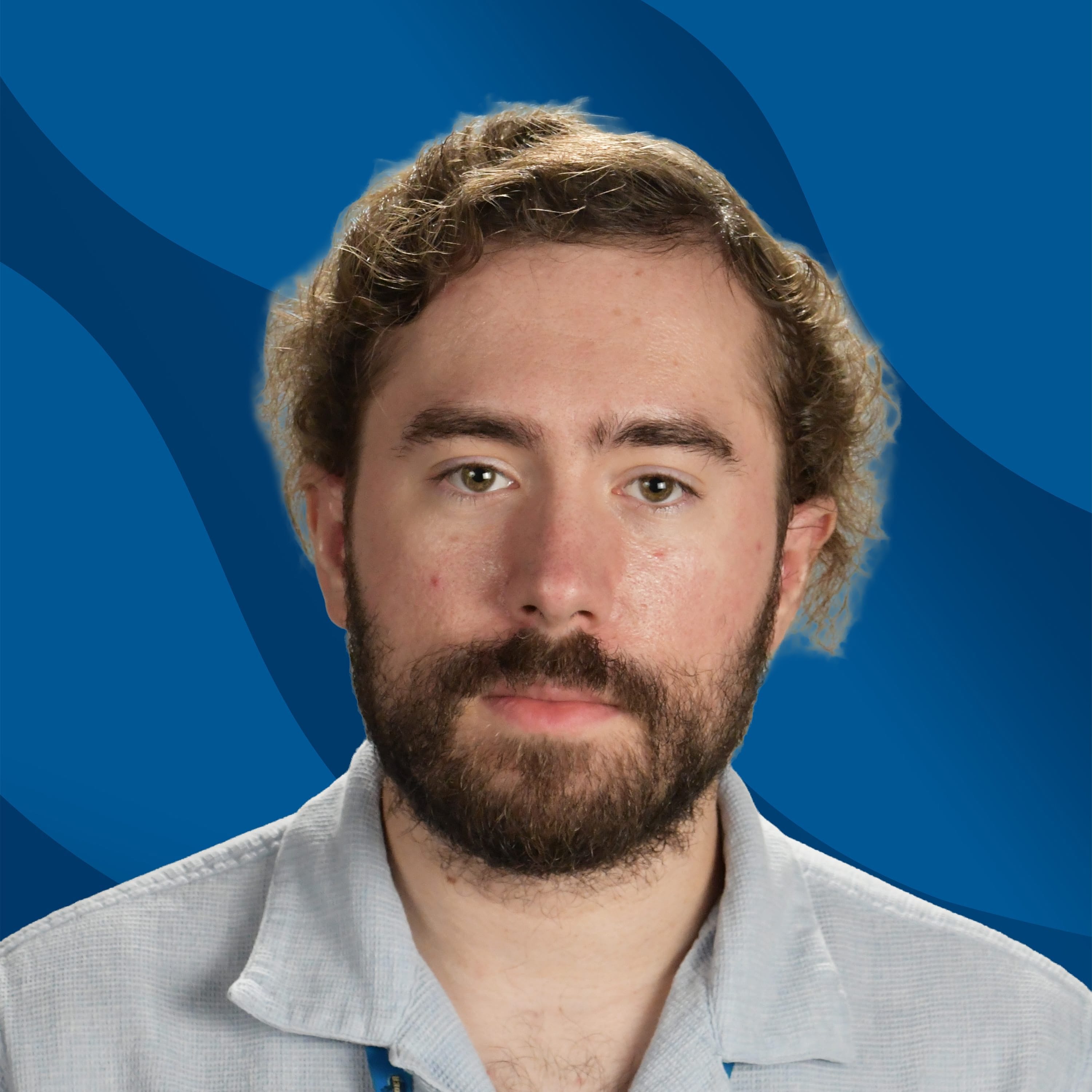A man with curly hair and a beard is wearing a light gray shirt, a blue lanyard, and a name tag, posing in front of a blue patterned background.