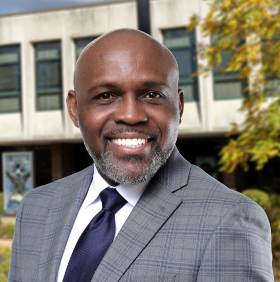 A smiling man in a grey plaid suit and navy tie stands outdoors in front of a modern building with large windows and autumn foliage.