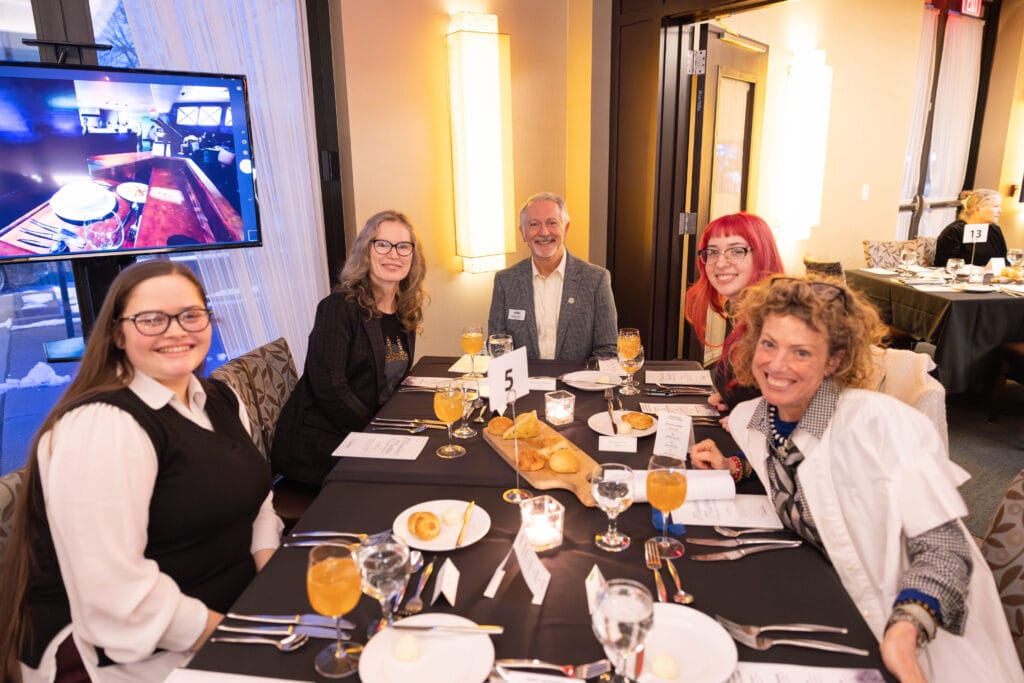 Five people sit around a round table set with food and drinks at a formal event. Everyone is smiling. A TV screen and other tables are visible in the background.