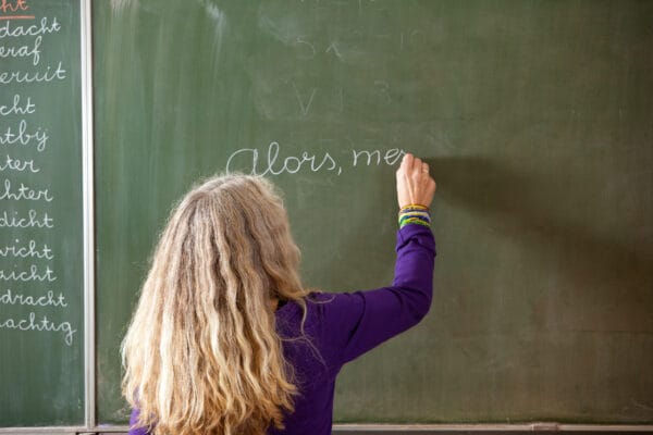 A teacher writing the words Alors, me in cursive on a green chalkboard.