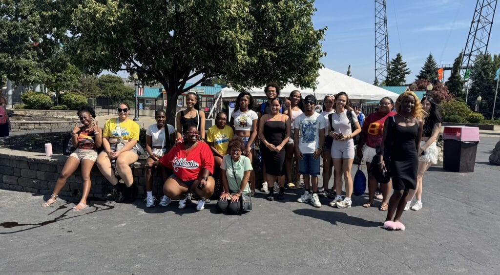 A large group of young adults posing for a group photo in front of a tree and a white canopy tent at an amusement park.