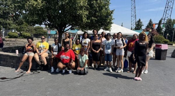 A large group of young adults posing for a group photo in front of a tree and a white canopy tent at an amusement park.