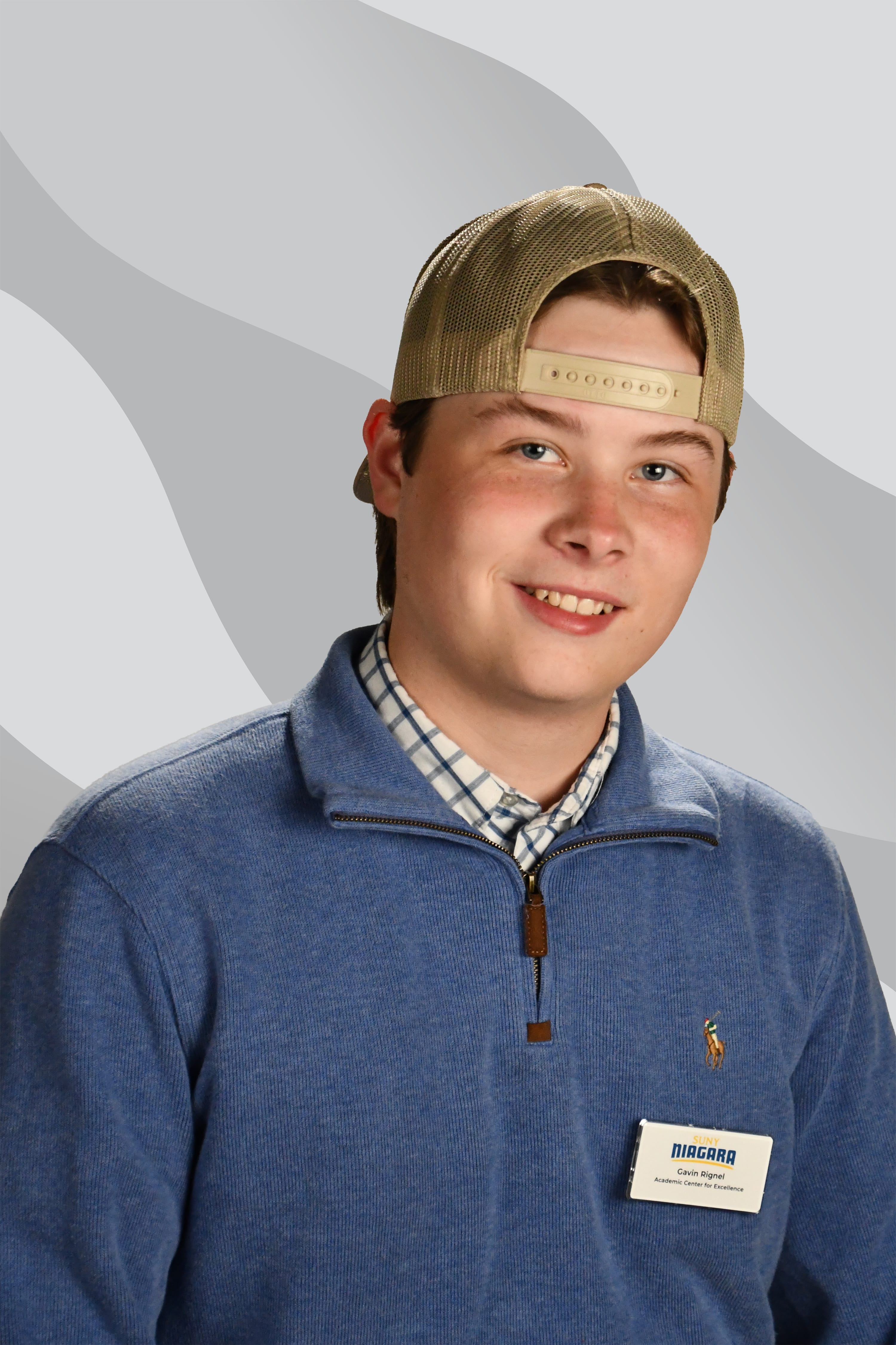 A young man smiling while wearing a mesh-back baseball cap and a blue quarter-zip sweater with a SUNY Niagara name tag.