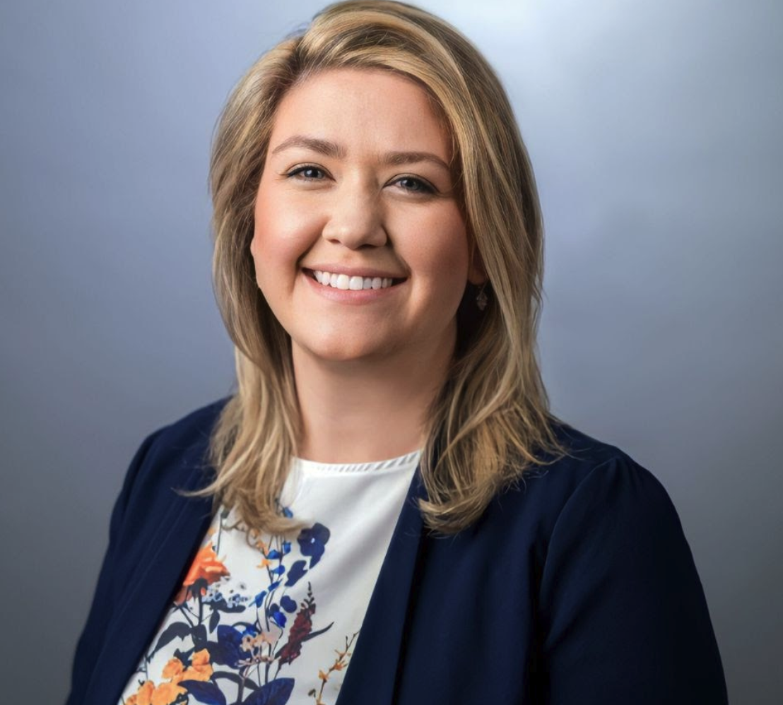 Smiling woman with blonde hair wearing a navy blazer and a white blouse with a colorful floral pattern, posed in front of a plain gray background.