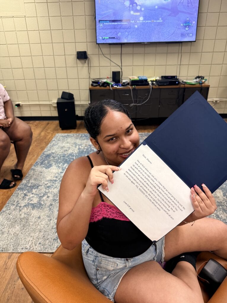 A smiling woman holds open a navy blue certificate folder, displaying a printed letter addressed to Sharon.
