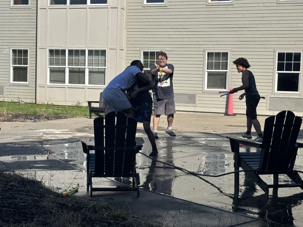 Four young adults playing with water guns on a wet patio outside an apartment building.