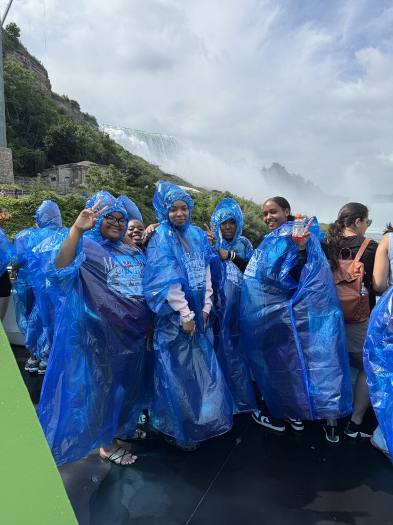 Five young women wearing blue plastic ponchos smile for a photo in front of Niagara Falls.