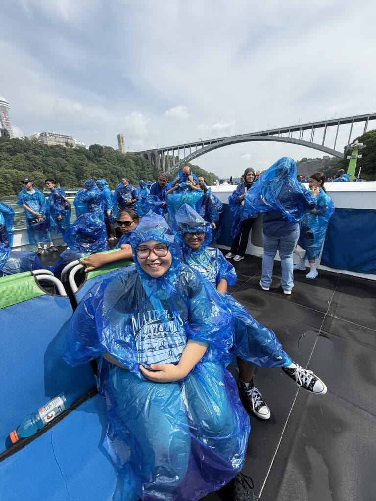 People wearing blue plastic ponchos on a boat deck in front of a large arch bridge.
