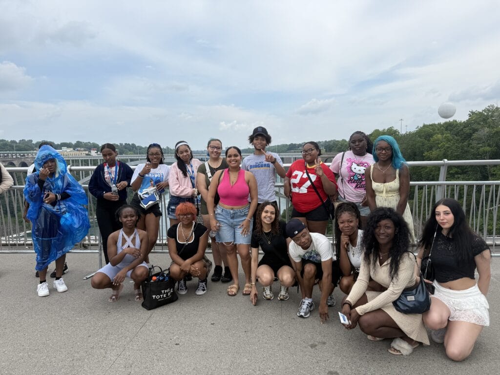 A large group of young adults posing for a photo on a pedestrian bridge overlooking Niagara Falls.