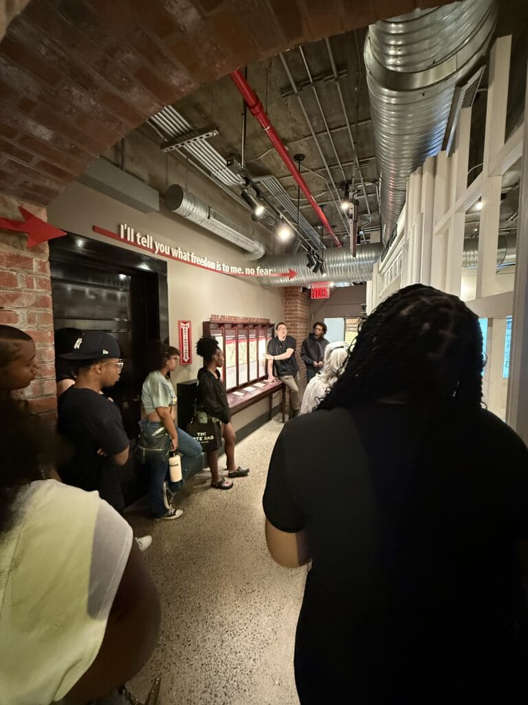 A group of people listens to a man speaking in a hallway with brick arches and a sign that reads Ill tell you what freedom is to me. no fear.