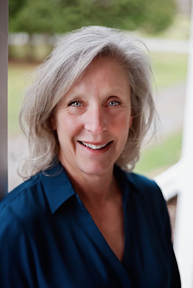 A smiling woman with shoulder-length grey hair wearing a teal shirt.
