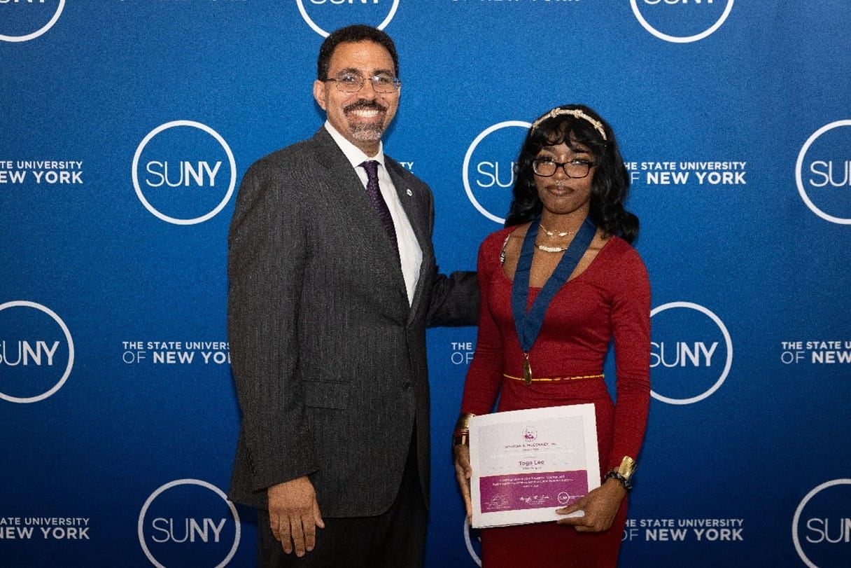 A man in a suit and a woman in a red dress stand in front of a SUNY branded backdrop; the woman holds a certificate and wears a medal.
